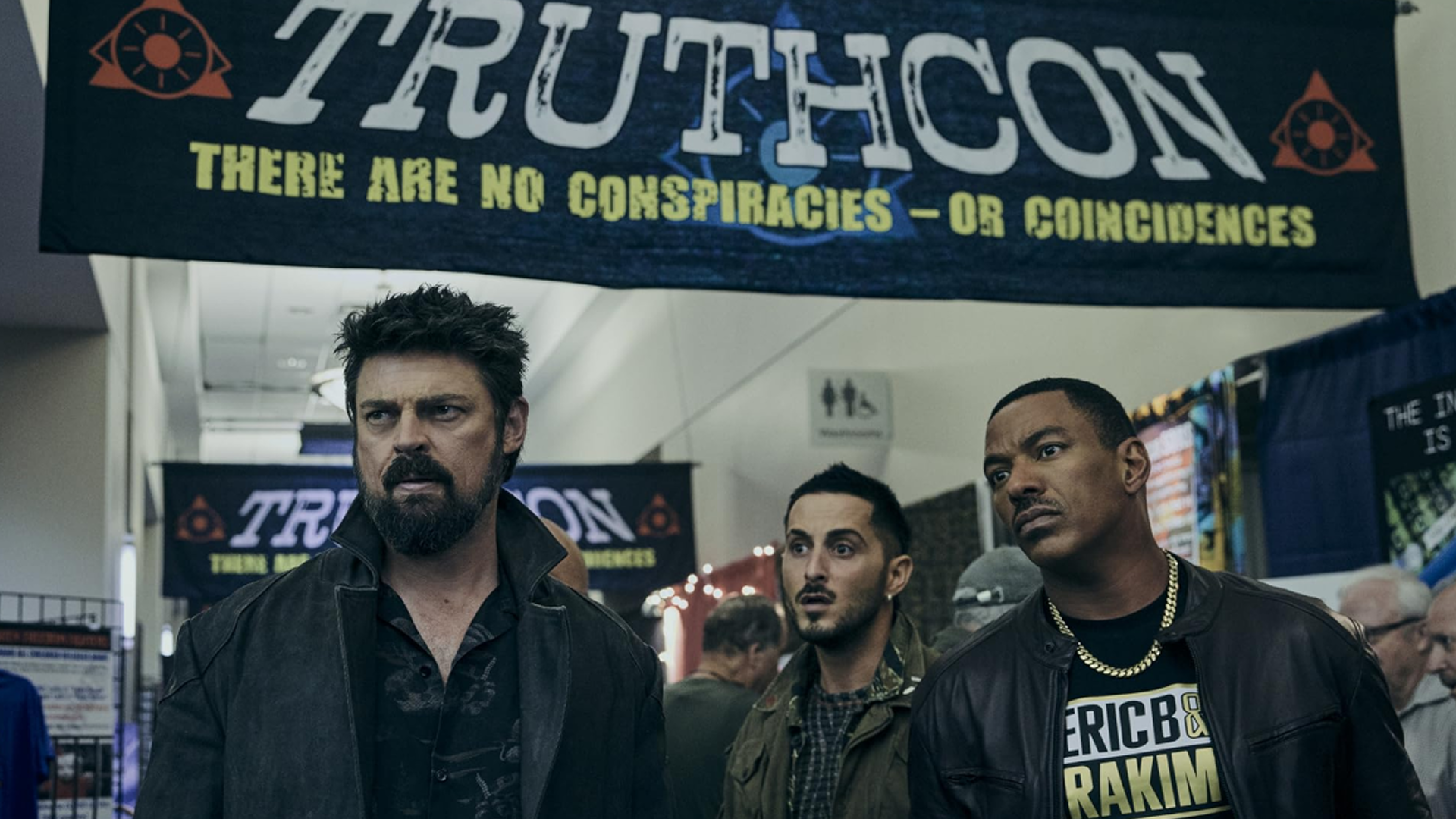 Billy Butcher, Frenchie, and Marvin Milk attend Truthcon in an episode of The Boys. The banner reads "Truthcon: There are no conspiracies - or coincidences"