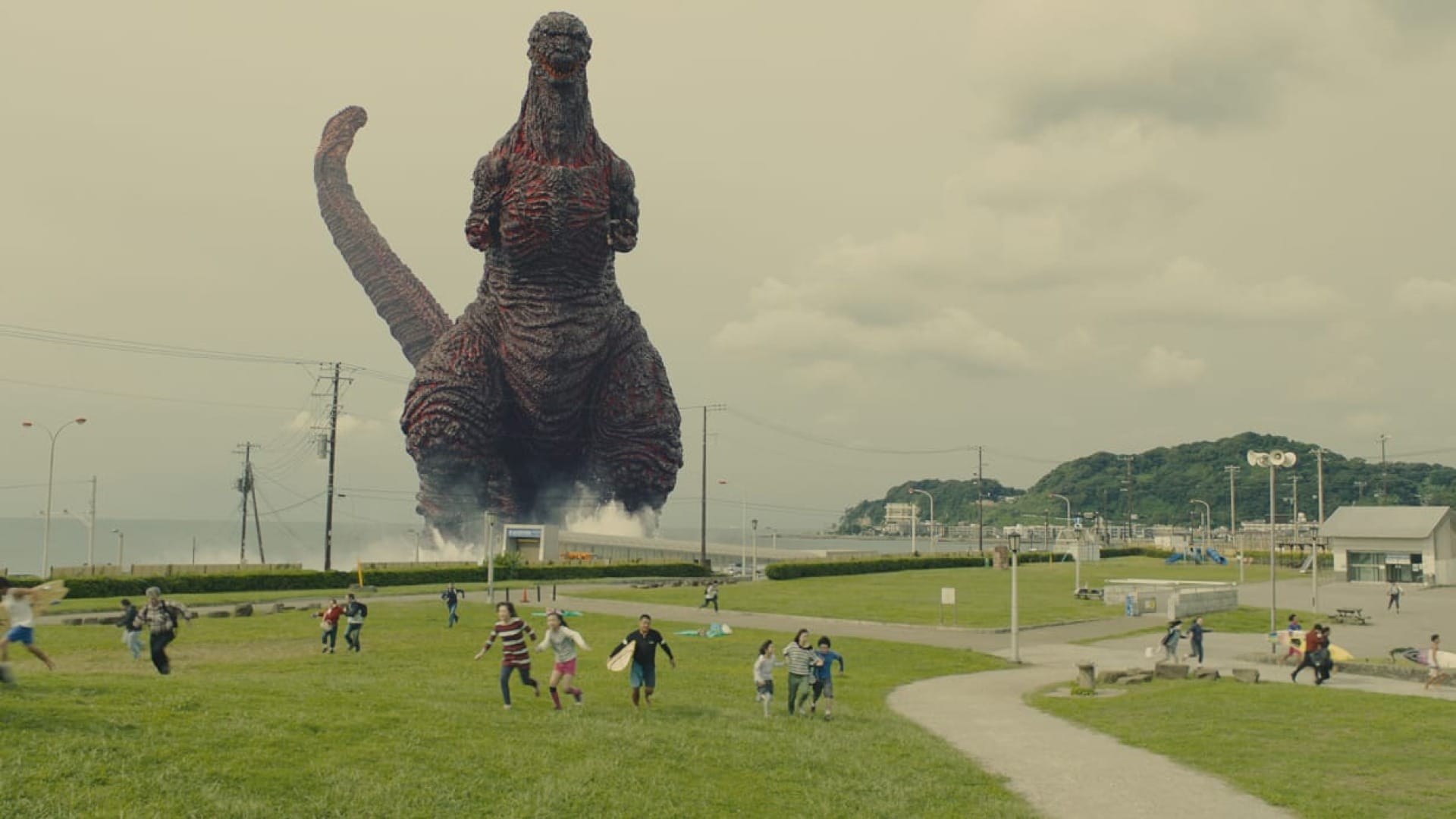 An image from Shin Godzilla (2016) with Godzilla walking ashore in the background while people run in the foreground.